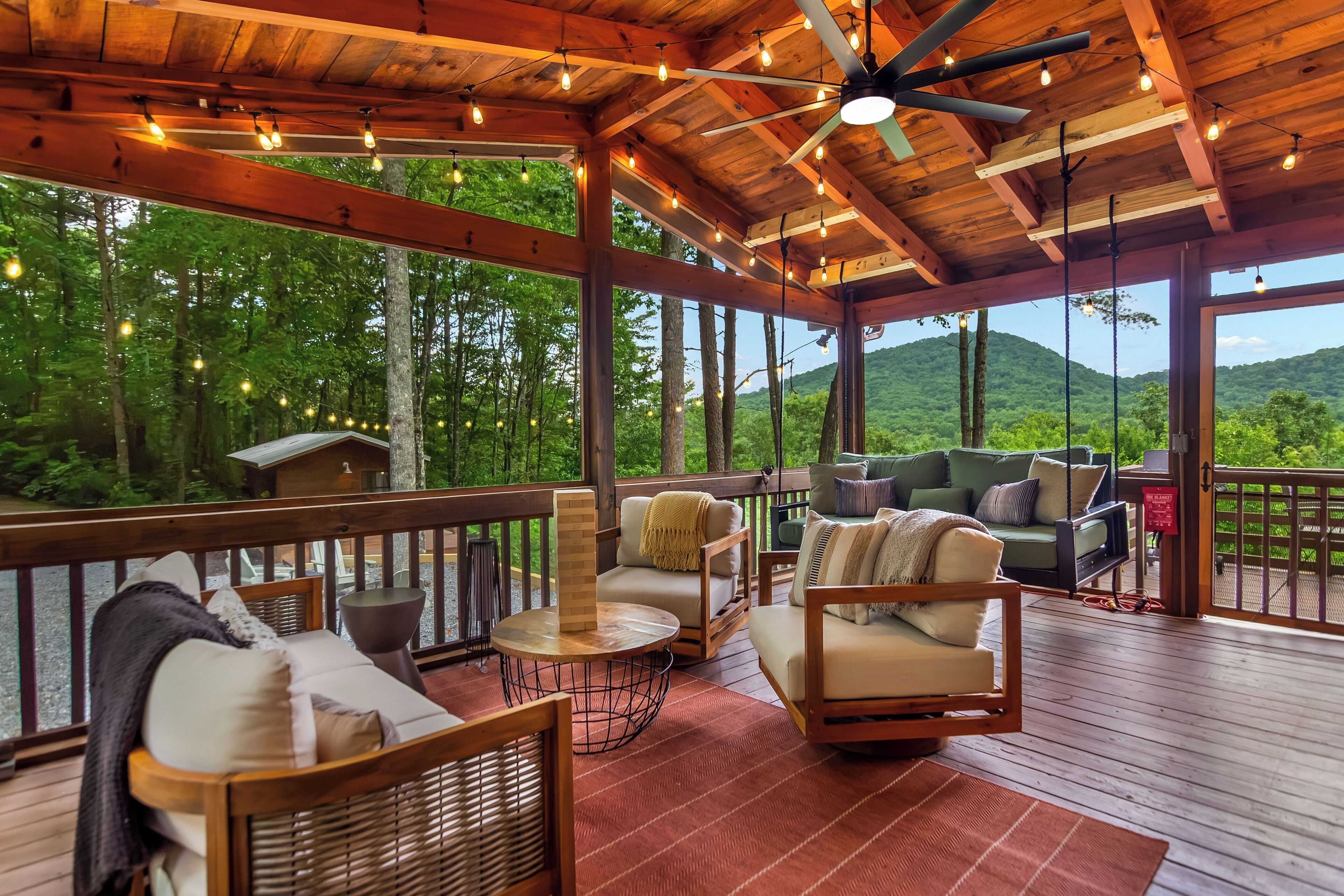A covered outdoor seating area with wooden furniture and string lights, overlooking a forested landscape.