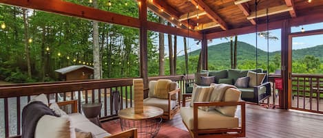 A covered outdoor seating area with wooden furniture and string lights, overlooking a forested landscape.