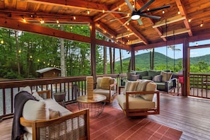 A covered outdoor seating area with wooden furniture and string lights, overlooking a forested landscape. - Luxe Mtn View 5 Acres, Hot Tub, Screened Porch (Mineral Bluff)