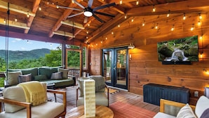 A wooden deck with a ceiling fan, string lights, a TV, and a green sofa with cushions. - Luxe Mtn View 5 Acres, Hot Tub, Screened Porch (Mineral Bluff)