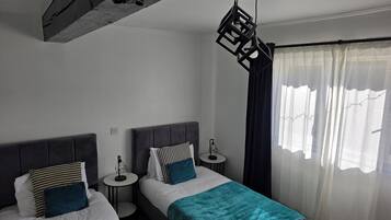 A bedroom with two beds, a small table, a pendant light, and a window with curtains.