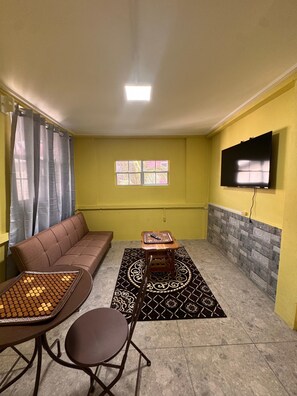 Uma sala de estar com sofá, uma mesinha com um tapete, televisão na parede e janela com cortinas.