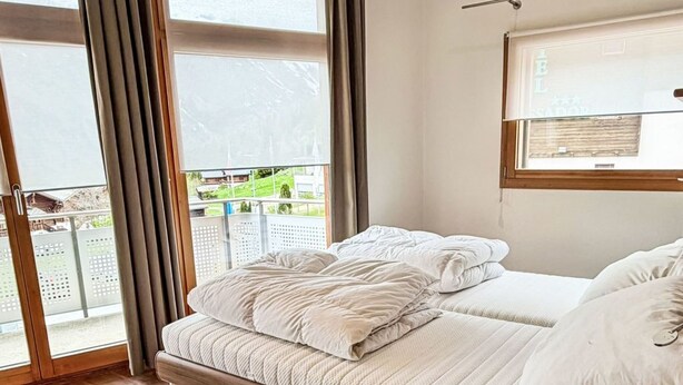 A bedroom with a wooden bed, large windows, and a view of the outdoors. - Haus zum Alpenblick - Wannenhorn by Interhome (Bellwald)