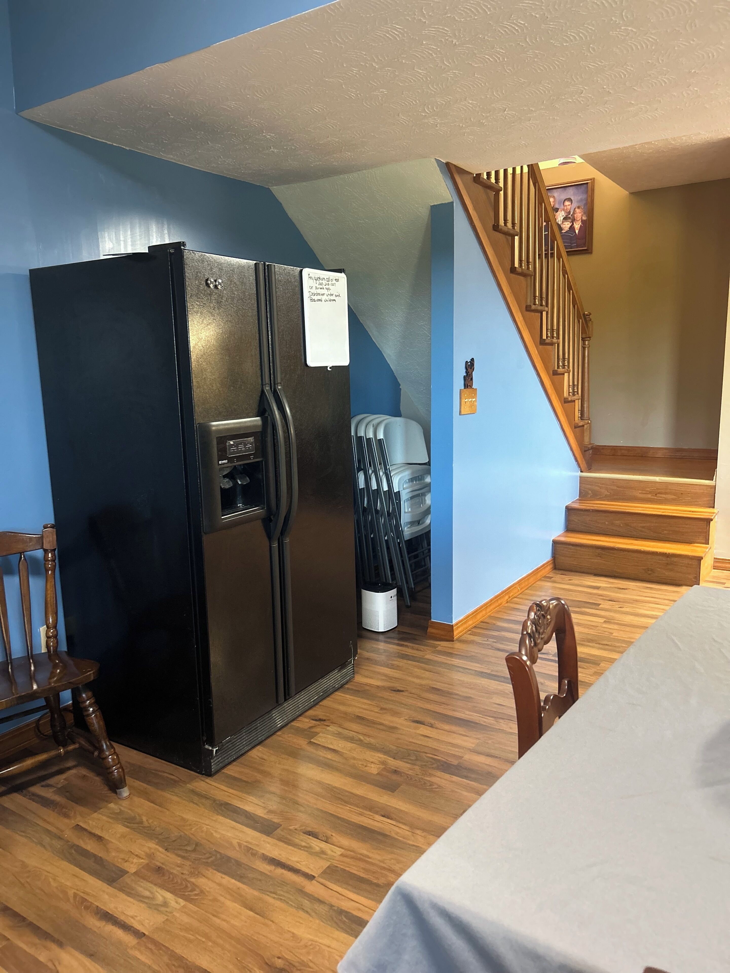 Una cocina con un refrigerador negro, sillas de madera, una mesa de comedor con mantel blanco y una escalera que sube a otro nivel.