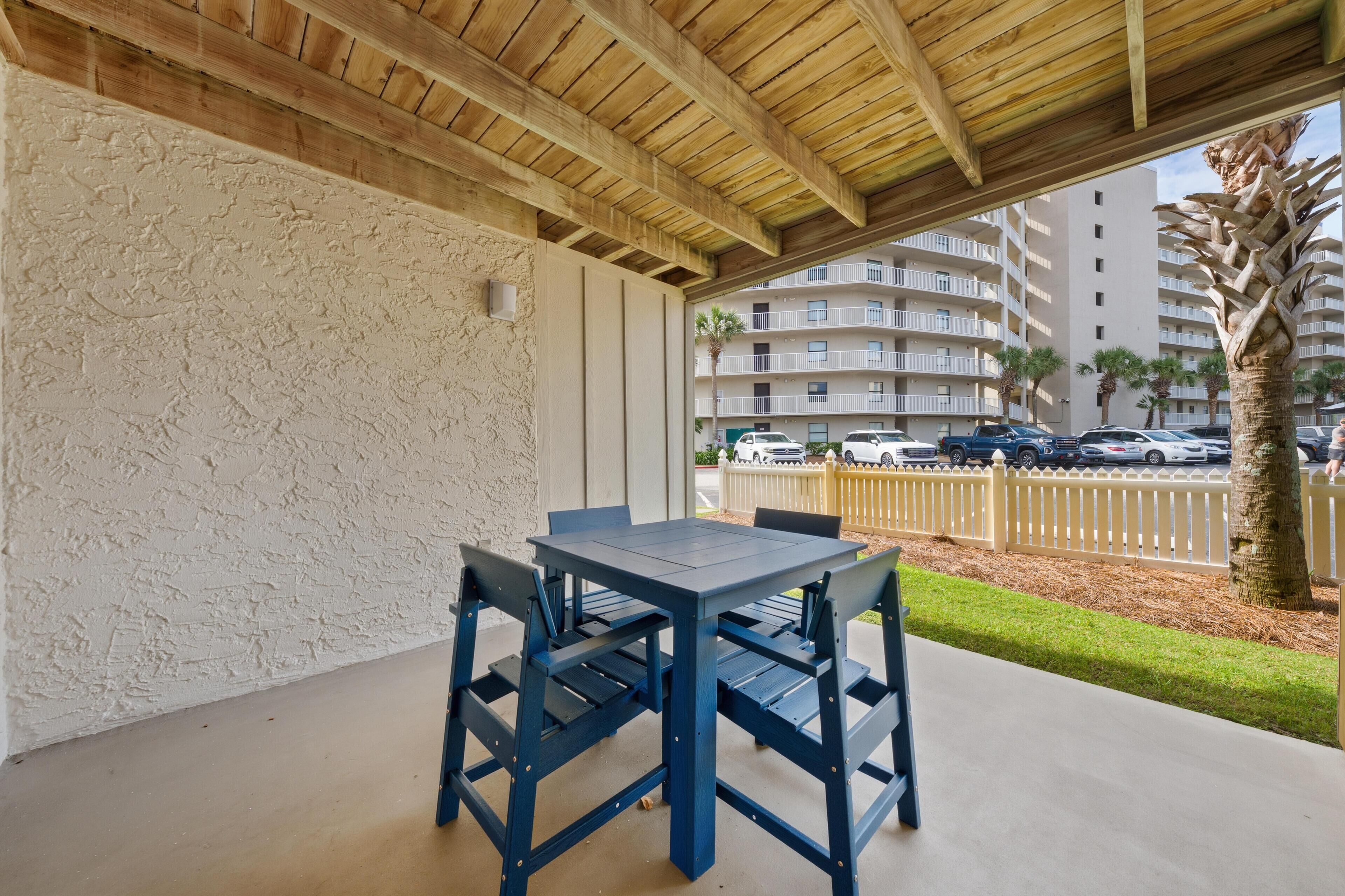 An outdoor patio with a table and chairs, a wooden ceiling, and a view of a multi-story building with parked cars and palm trees.