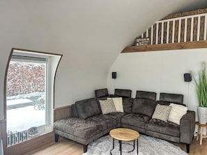 Een moderne woonkamer met een hoekbank, een ronde salontafel en een groot raam met uitzicht op een besneeerd landschap.