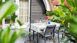 Un patio exterior con una mesa lista para dos personas, una botella de vino blanco y un florero con flores.