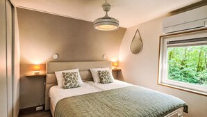Un dormitorio con cama, lámparas de noche, ventilador de techo, ventana con vista a follaje y mesita de noche.