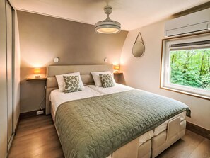 Quarto com cama, luminárias de cabeceira, ventilador de teto, janela com vista para o verde e mesa de cabeceira.