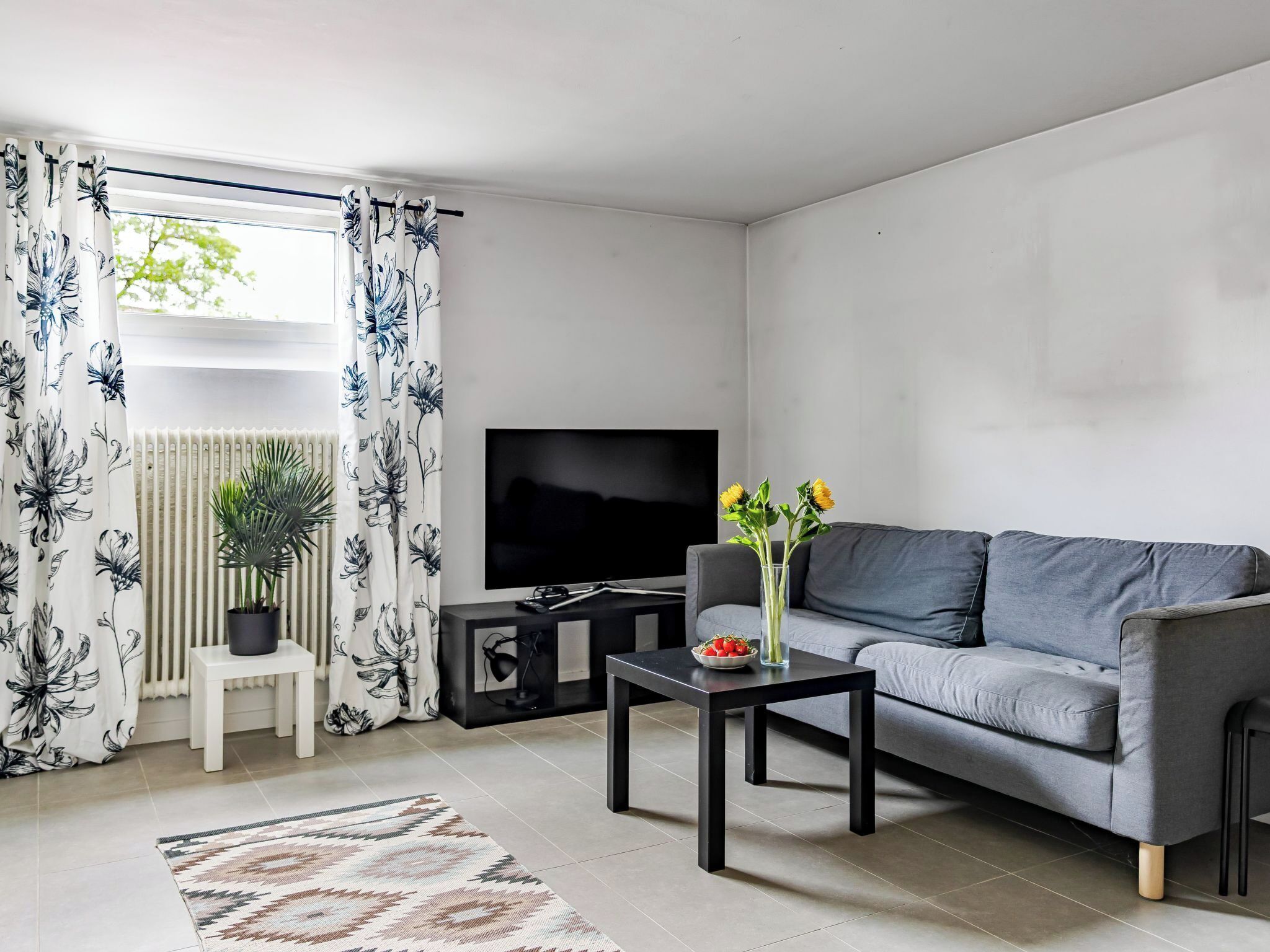 Een moderne woonkamer met een grijze bank, een flatscreen televisie, een tafeltje met een bloevas, een radiator en een raam met bloemen gordijnen.