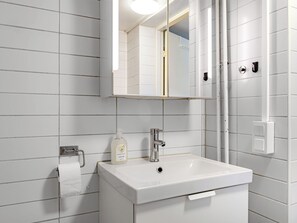 Un baño con paredes de azulejos blancos, un lavamanos blanco, un espejo, un dispensador de jabón y un perchero para toallas.