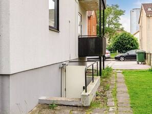 En bostadsbyggnad med vit fasad, en liten uteplats med en gång av sten, en parkerad bil och en grön soptunna.
