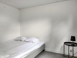 Una cama individual con ropa blanca, en una habitación con paredes blancas lisas y una pequeña mesa negra con una lámpara.