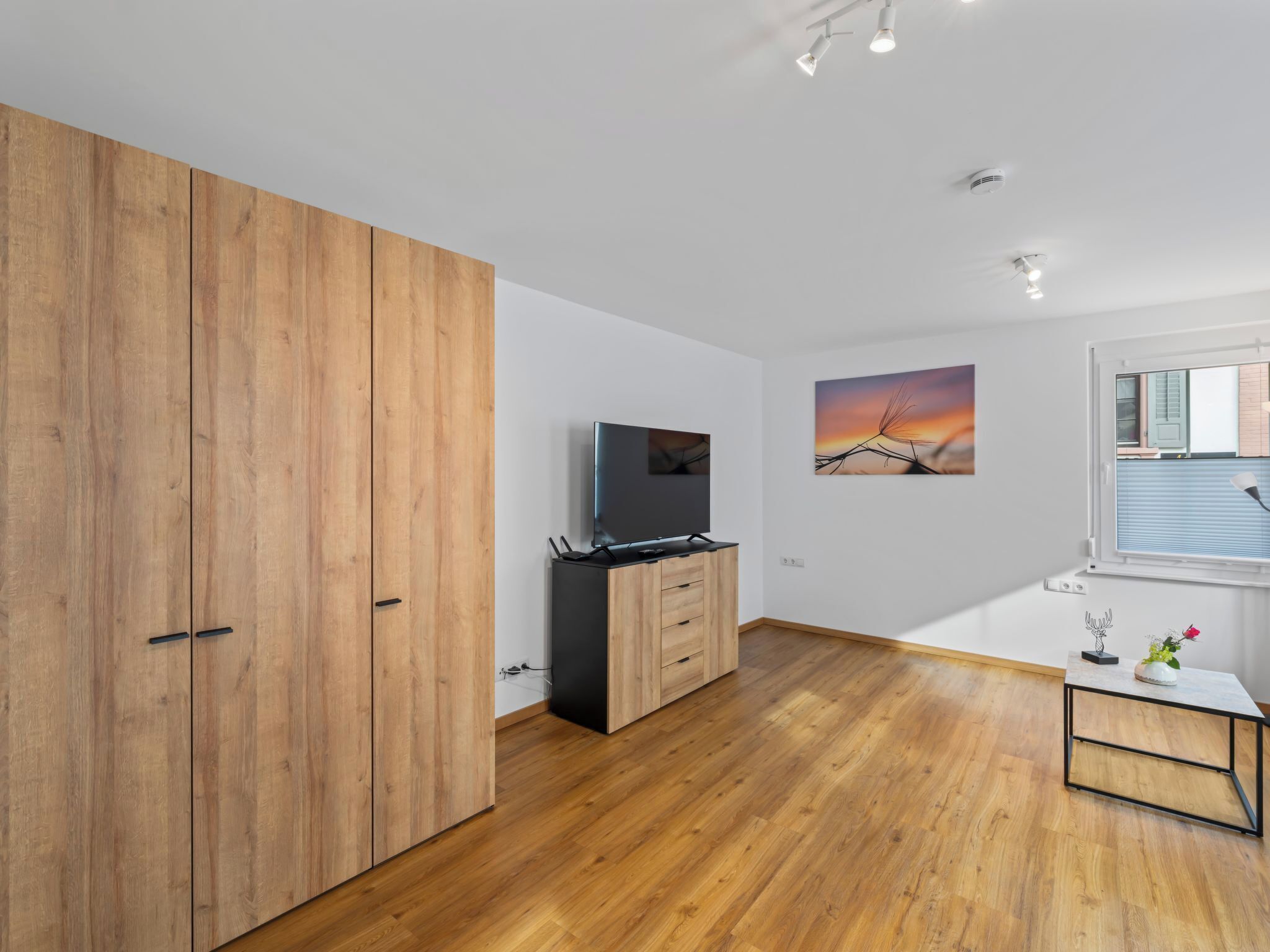 Ein modernes Zimmer mit Holzboden, einem Fernseher, einem Schrank, einem Gemälde und einem Fenster mit Jalousien.