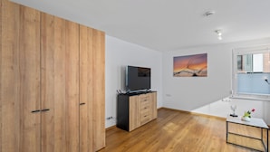 Ein modernes Zimmer mit Holzboden, einem Fernseher, einem Schrank, einem Gemälde und einem Fenster mit Jalousien.