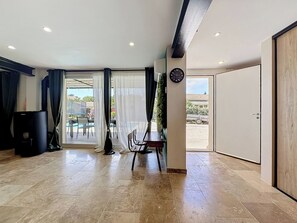 A spacious room with a tiled floor, a dining area with a table and chairs, a black heater, and a large window with curtains. - La Villa Soleya by Interhome (Rodilhan)