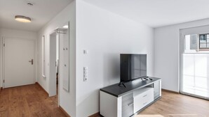 Ein modernes Wohnzimmer mit Flachbildfernseher auf einer Kommode, einer Tür zu einem anderen Raum und einem Fenster mit Jalousien.