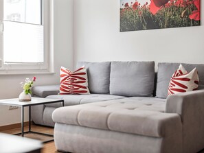 A modern living room with a grey sectional sofa, a small white side table, and a vase with flowers. - Birgit (S2) by Interhome (Endingen am Kaiserstuhl)