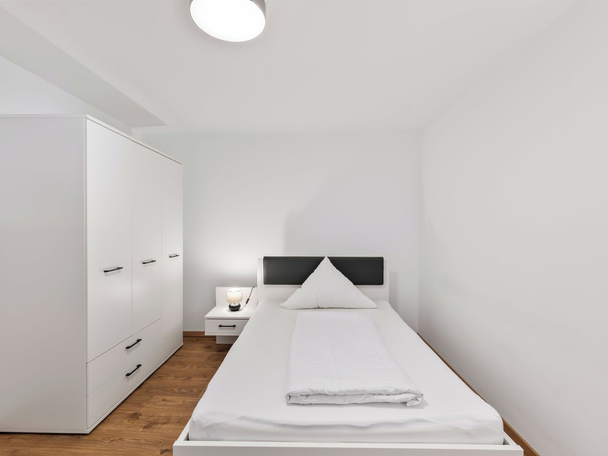 Un dormitorio minimalista con una cama individual, una mesita de noche, un armario y una lámpara de techo.