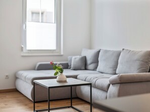 Ein modernes Wohnzimmer mit einem grauen Sofa, einem kleinen schwarzen Metalltisch mit einer weißen Vase und einem Fenster mit Jalousien.