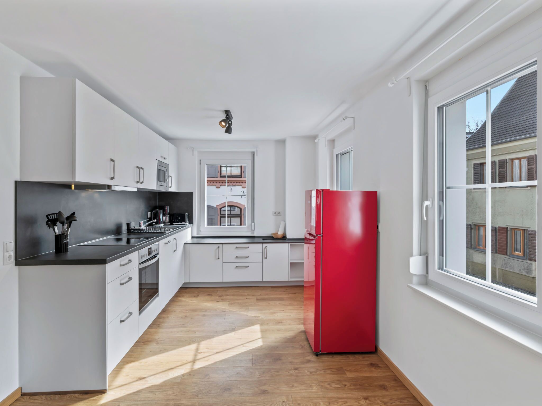Une cuisine moderne avec des meubles blancs, un plan de travail noir, un réfrigérateur rouge et une fenêtre donnant sur des immeubles.