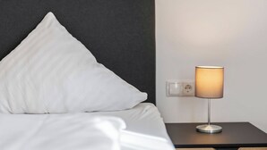 A neatly made bed with white linens and a dark headboard, a bedside table with a lamp, and a power outlet on the wall. - Diana (S4) by Interhome (Endingen am Kaiserstuhl)