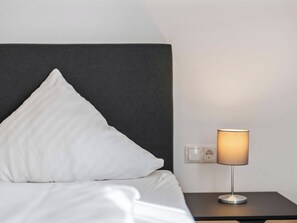 Ein ordentlich bezogenes Bett mit weißen Laken und einem dunklen Kopfteil, ein Nachttisch mit Lampe sowie eine Steckdose an der Wand.