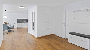 Ein moderner, gut beleuchteter Raum mit Holzboden, weißen Wänden, einem Essbereich mit Tisch und Stühlen sowie einer Küche mit weißen Schränken und einer Sitzbank mit Staufächern.