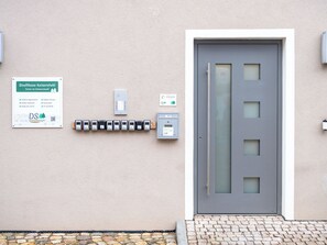 Ein Gebäudeeingang mit einer grauen Tür, einem Sicherheitstastenfeld und einem Schild, das Annehmlichkeiten wie moderne Apartments, Balkone sowie Fernsehen und WLAN auflistet.