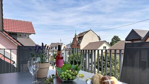Ein Balkon mit Tisch, gedeckt für eine Mahlzeit, mit einer Flasche, einer Schüssel mit Trauben, einer Brotteller, einem Glas und einer Zeitschrift.