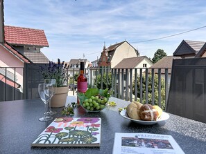Ein Balkon mit Tisch, gedeckt für eine Mahlzeit, mit einer Flasche, einer Schüssel mit Trauben, einer Brotteller, einem Glas und einer Zeitschrift.