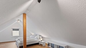 Ein modernes, helles Schlafzimmer im Dachgeschoss mit schrägem Dach, einem Holzbalken, einem Bett, Nachttischen und integrierten Regalen.