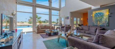 A spacious living room with large windows, a sofa set, a coffee table, and a painting on the wall.