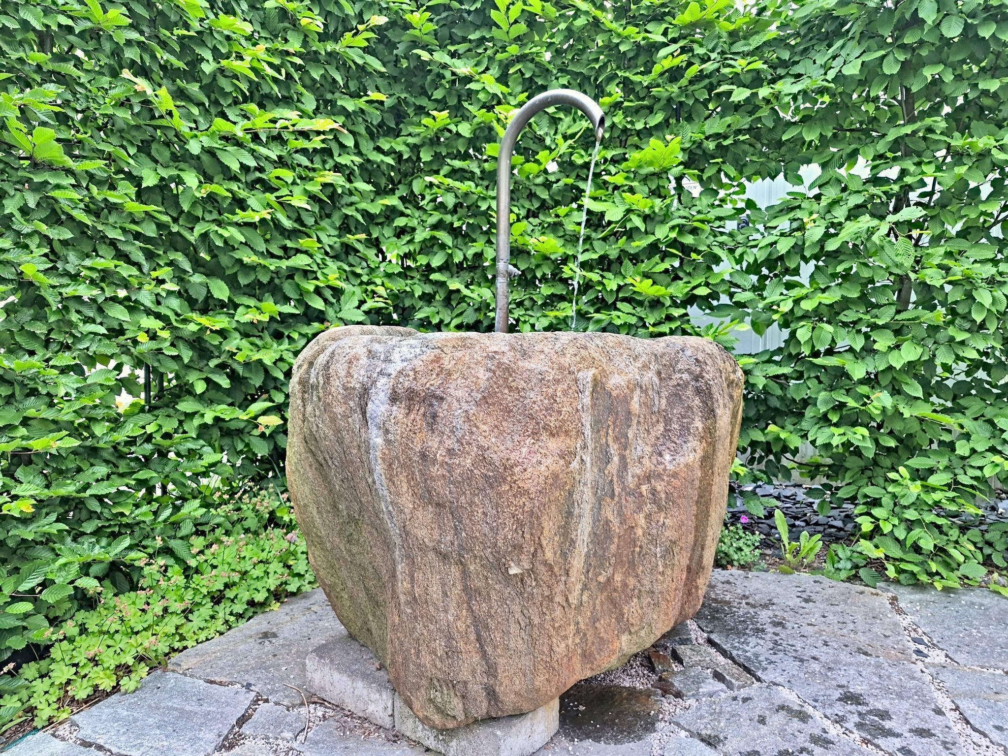 Una gran estructura de pedra erosionada amb un broc metàl·lic corbat, situada sobre una base de pedra, envoltada d'una vegetació verda densa.