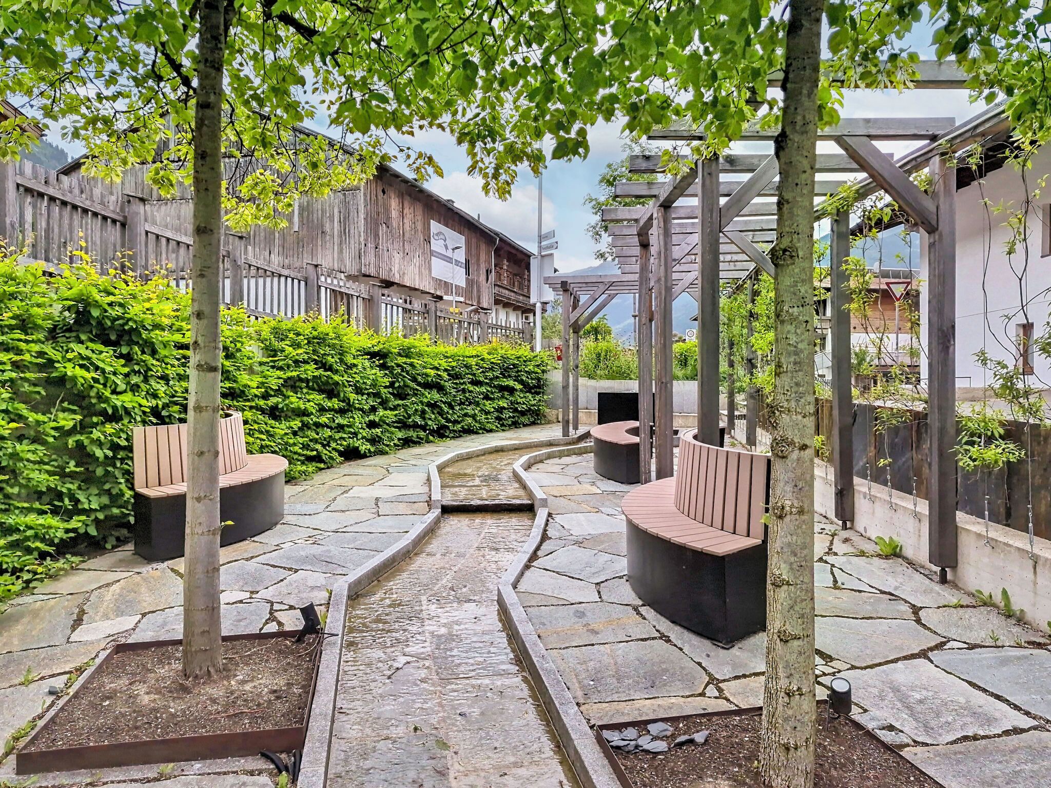 A stone pathway leading to a seating area with benches, surrounded by lush greenery and a wooden fence.