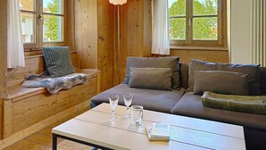 Una habitación acogedora con un banco de madera, un sofá, una mesa con vasos y un libro, y ventanas amplias con cortinas.