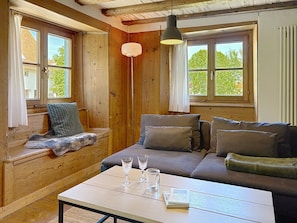 Ein gemütliches Zimmer mit einer hölzernen Bank, einem Sofa, einem Tisch mit Gläsern und einem Buch sowie großen Fenstern mit Vorhängen.