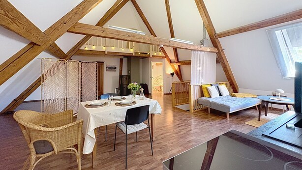 Een kamer in loftstijl met zichtbare houten balken, een eettafel met stoelen, een bank en een televisie.