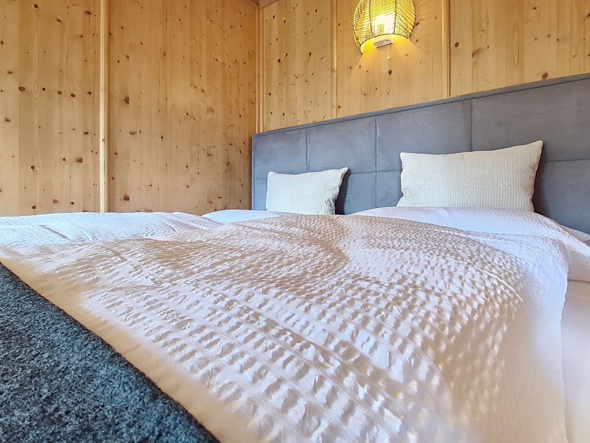 A bedroom with a wooden wall, a grey headboard, and a white bedspread.