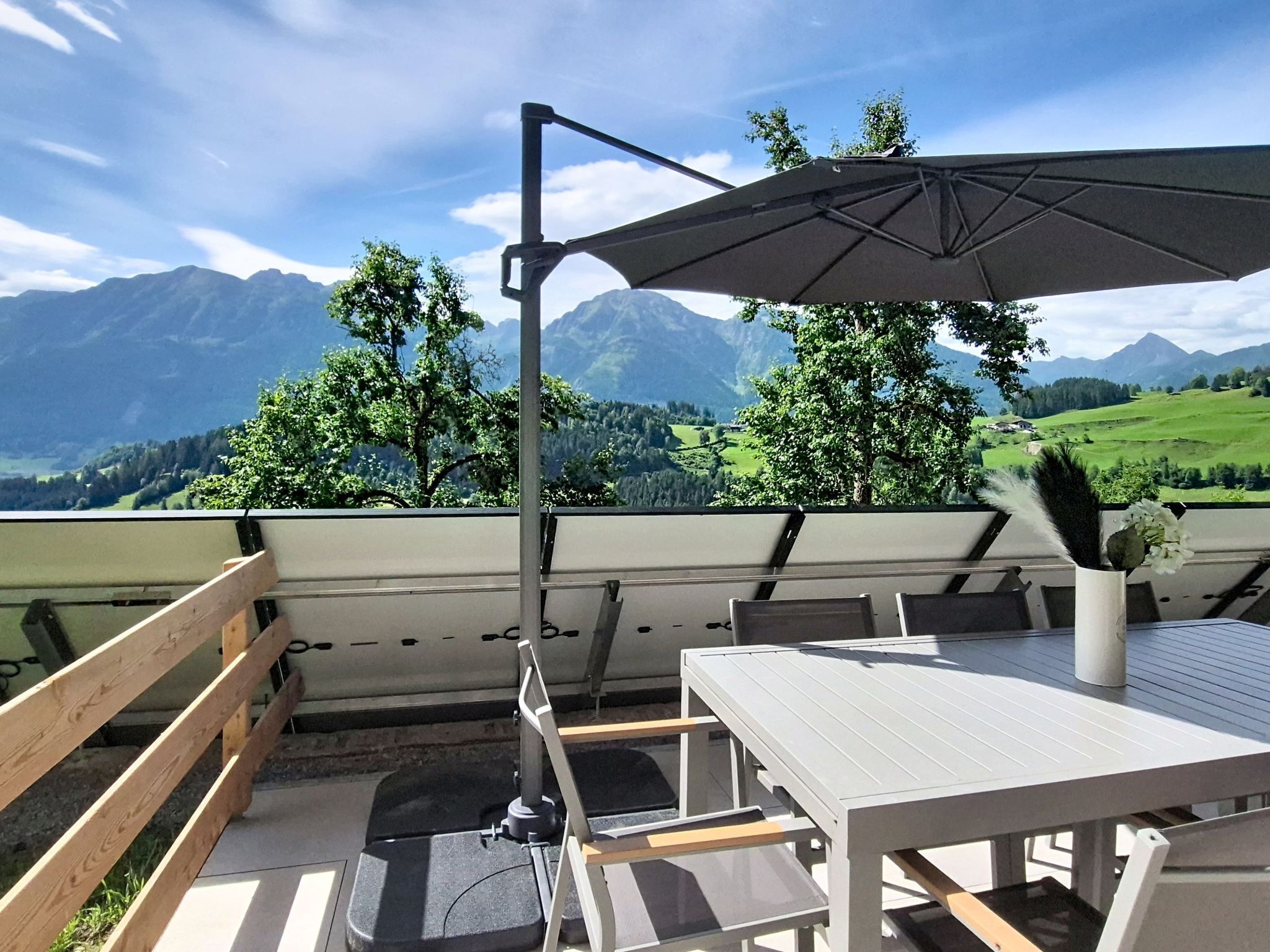 A terrace with a table and chairs, a wooden railing, and a view of mountains and greenery.