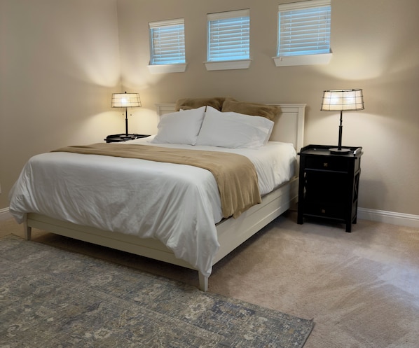 Une chambre à coucher comprenant un lit, deux tables de chevet, un tapis et trois fenêtres équipées de stores.