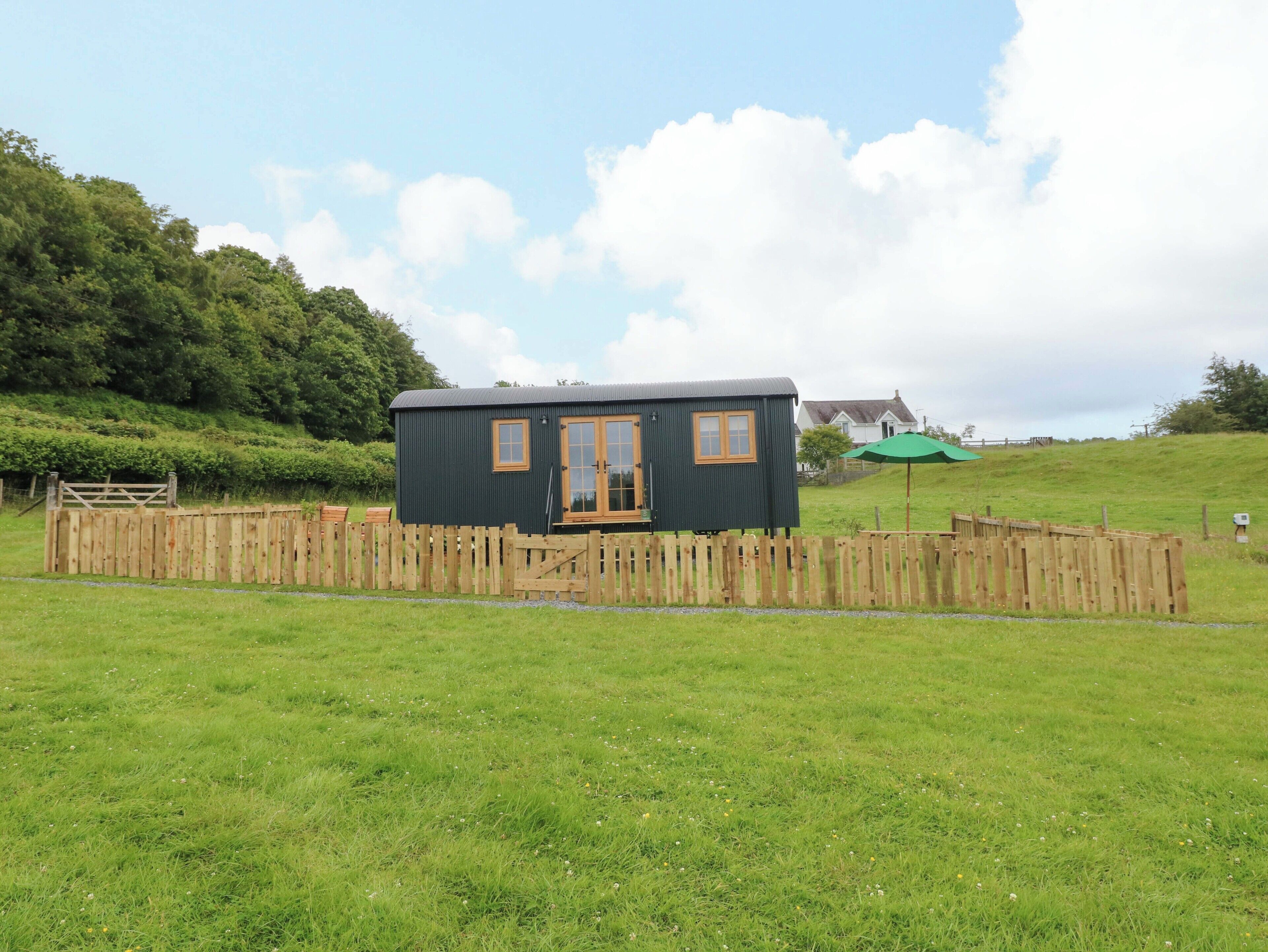 A small, dark-colored cabin with a wooden door and windows, surrounded by a wooden fence and a grassy field.