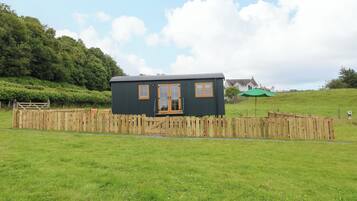 A small, dark-colored cabin with a wooden door and windows, surrounded by a wooden fence and a grassy field.