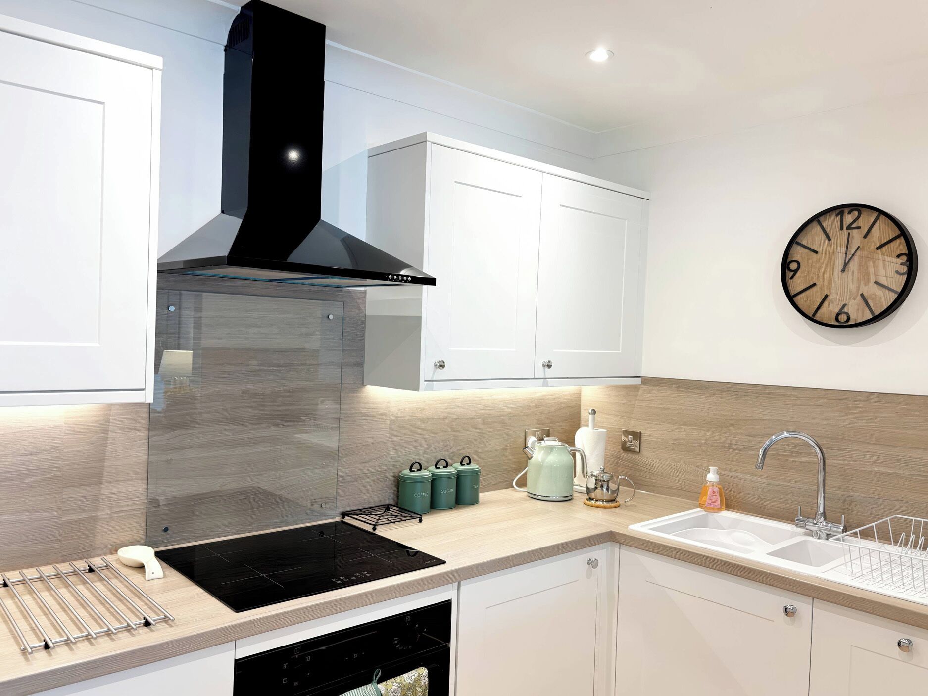 Una cocina moderna con armarios blancos, una campana extractora negra, horno empotrado, un fregadero y un reloj de pared.