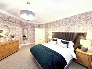 A bedroom with a large bed, a dresser, a nightstand with a lamp, a mirror, and a door leading to another room.