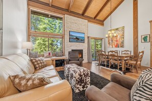 A cozy living room with a fireplace, large windows, a dining area, and a view of the outdoors. - Trailside Condo | Steps to Shuttle & Village (Truckee)