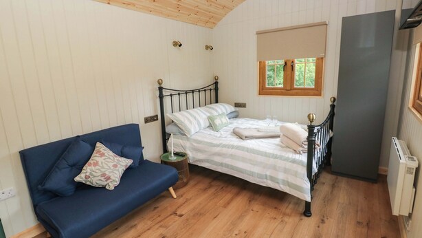 A bedroom with a bed, a blue sofa, a small bedside table, a window, and a radiator. - Cwtch Y Bugail 1 (Llandeilo)