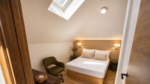 Quarto com claraboia, cama, cadeira, mesa de cabeceira e uma pequena planta.