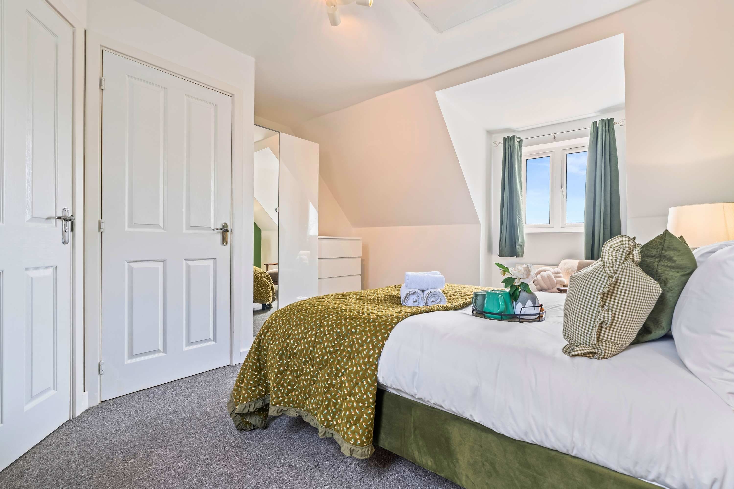 A hotel room with a bed, a window with curtains, a desk with a lamp, and a mirror.