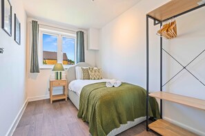 A bedroom with a bed, a bedside table, a lamp, a window with curtains, a radiator, and a wooden shelf. - 4 BR House with Private Garden, 2X FREE Parking (Basingstoke)